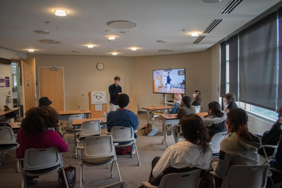 The image captures the student and staff in attendance. Omar Alshogre is pictured speaking in front of his audience. There is a monitor depicting those attending the event virtually.