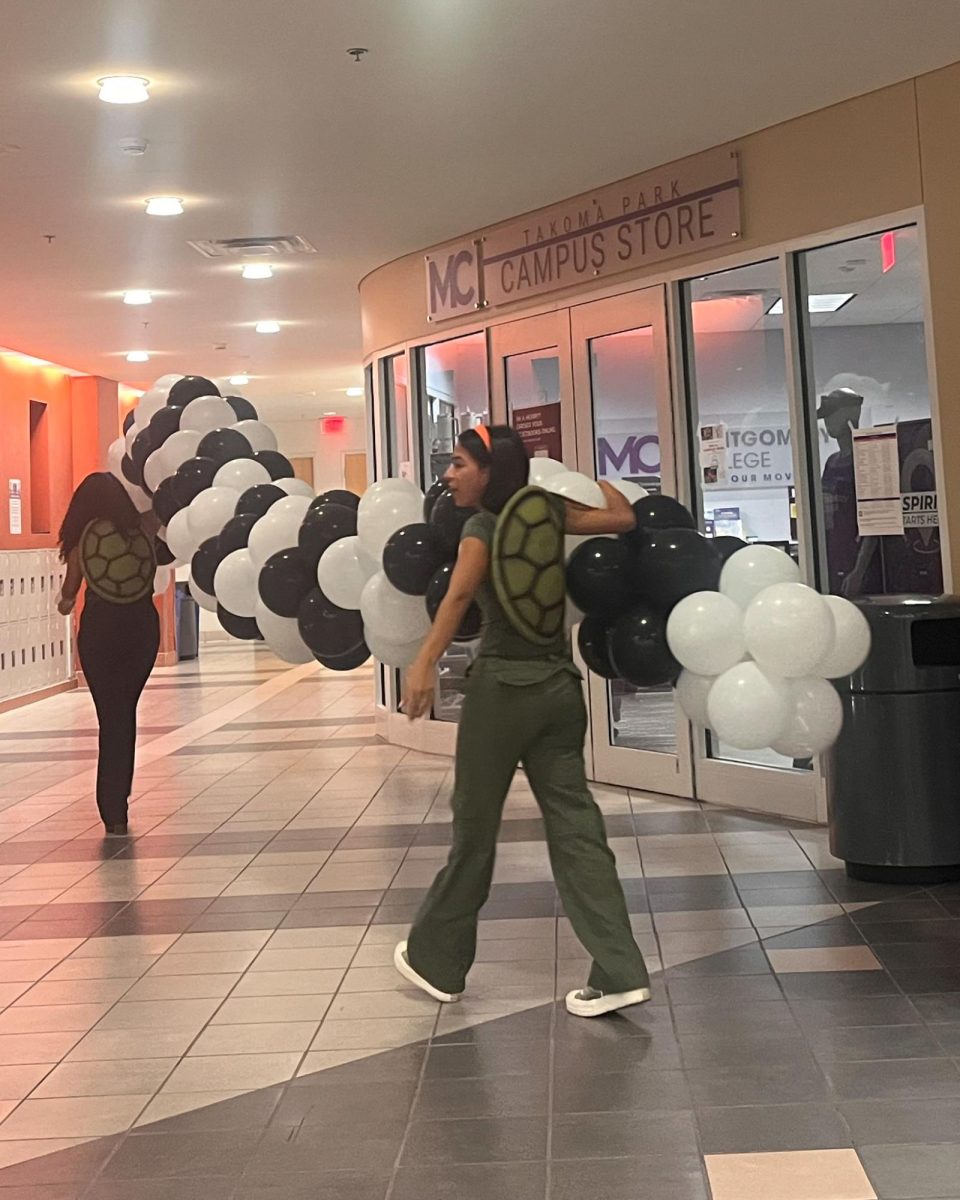 Sofia Perez, chair of the Student Activities Board, is helping Makeya Segu, a Pre-Clinical & General Studies: STEM student, carry a black and white balloon arch outside the Takoma Park Campus Store during the clean-up portion of the event. They are both wearing matching turtle costumes.